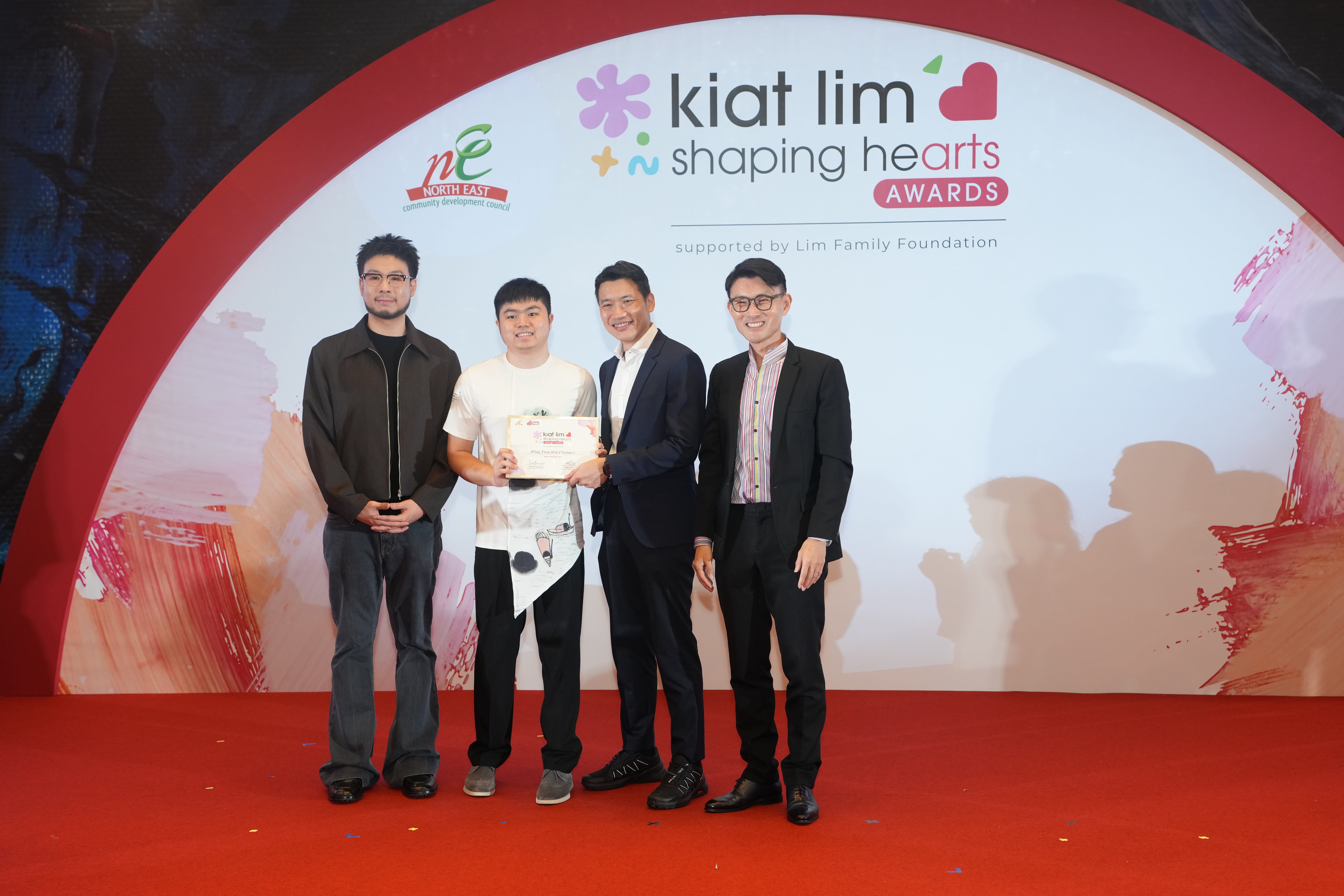 Award recipient standing on stage with the presenter, holding their certificate and posing for a group photo during the award ceremony, with a backdrop reading ‘Kiat Lim-Shaping Hearts Awards'.
